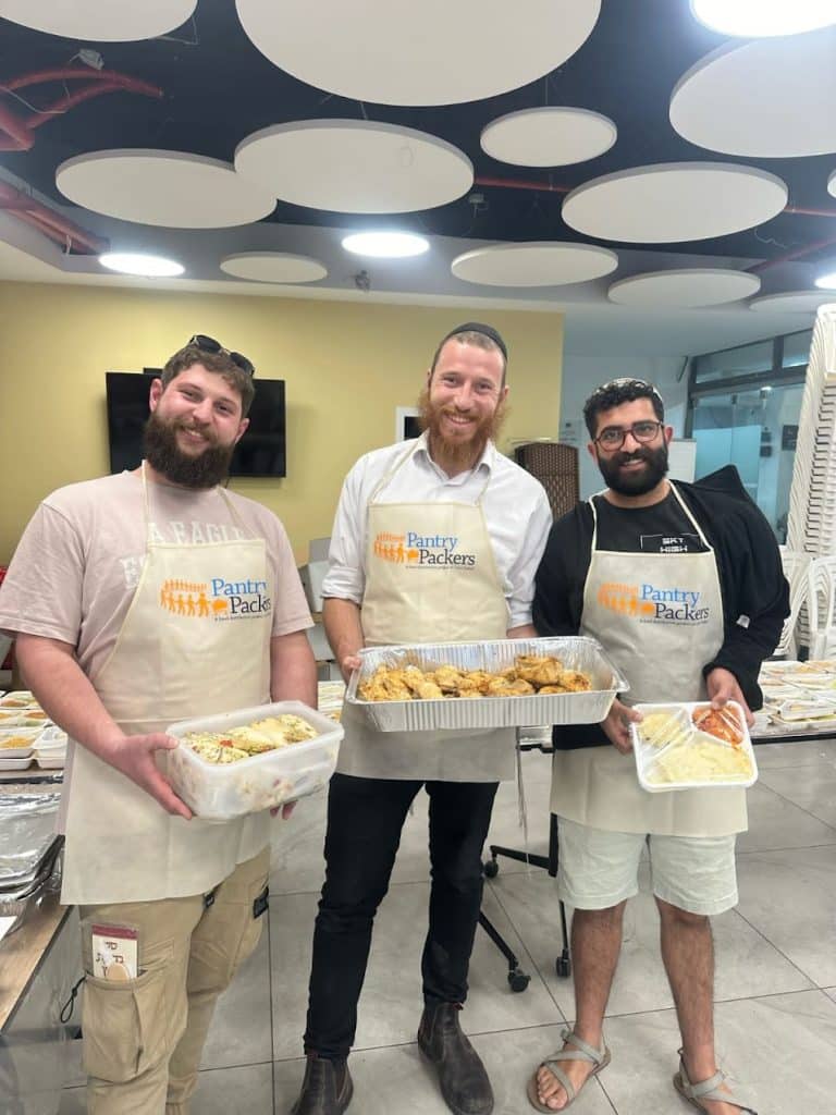 Trois hommes souriants portant des tabliers, chacun tenant un récipient contenant de la nourriture.