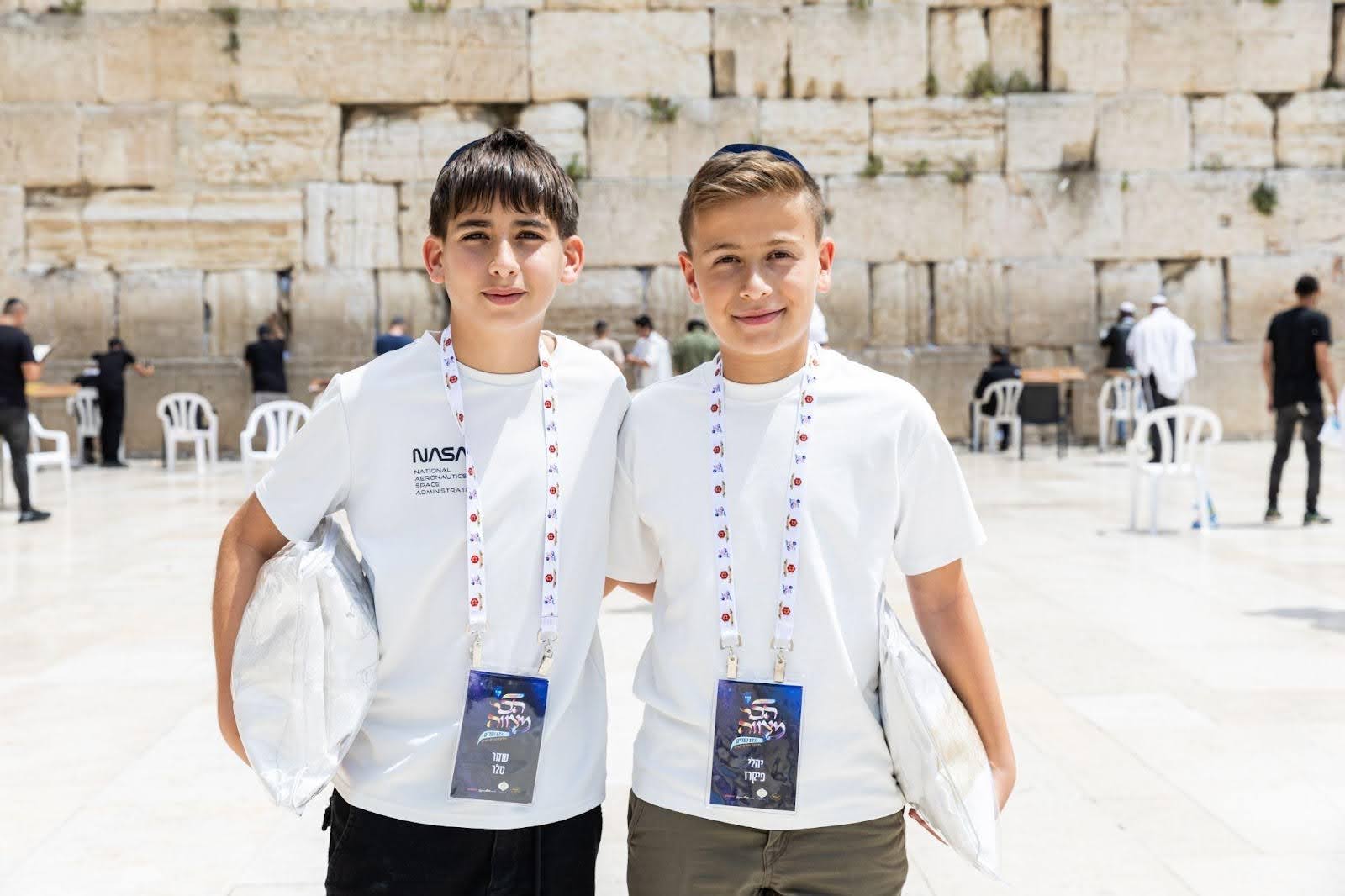 Two boys in white t-shirts and lanyards stand smiling at the Western Wall in Jerusalem, one wearing a NASA shirt, with prayer bags tucked under their arms.