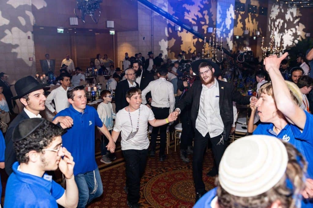 A joyful dancing chain of men and boys in yarmulkes, some with black hats, at a crowded banquet hall event with candelabras and patterned lighting.