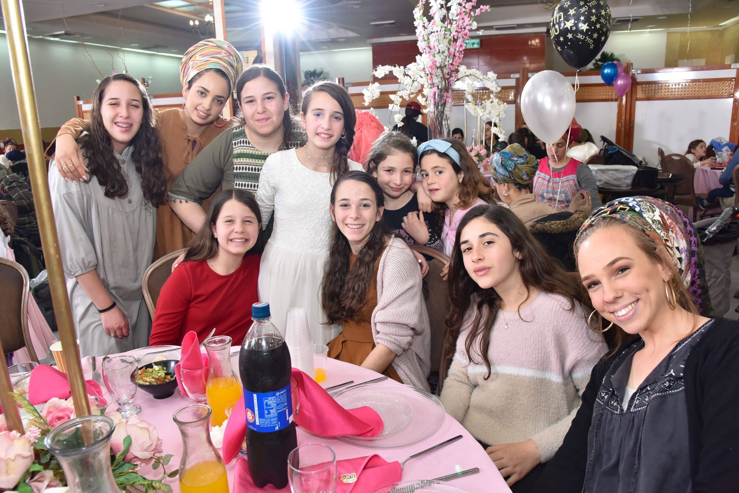A group of young women and girls gathered around a decorated table at a celebration, smiling for a photo. They are dressed in modest formal clothing, with some wearing head coverings. The table has pink napkins, flowers, drinks, and place settings, while balloons and floral decorations fill the background of the festive indoor venue.