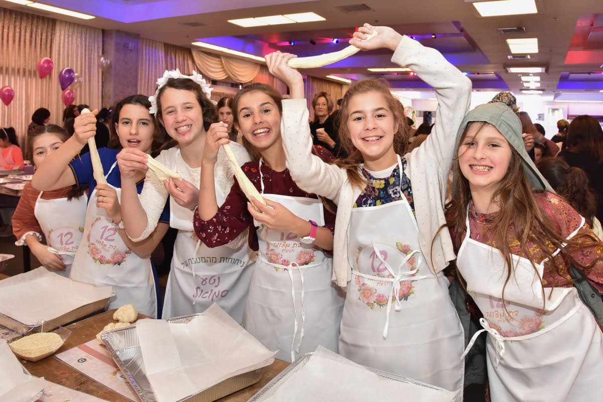 Girls participate in a community challah-baking event, stretching dough and smiling while gathered around a baking table.